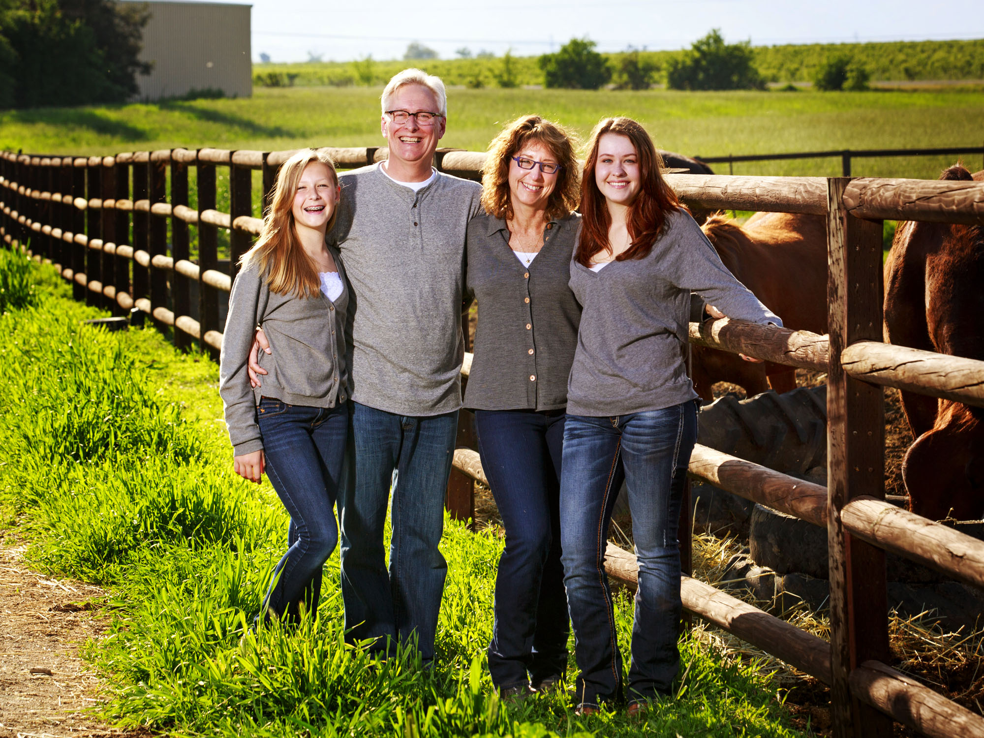 Family of four standing by a wooden fence with brown cows in a green field on a sunny day