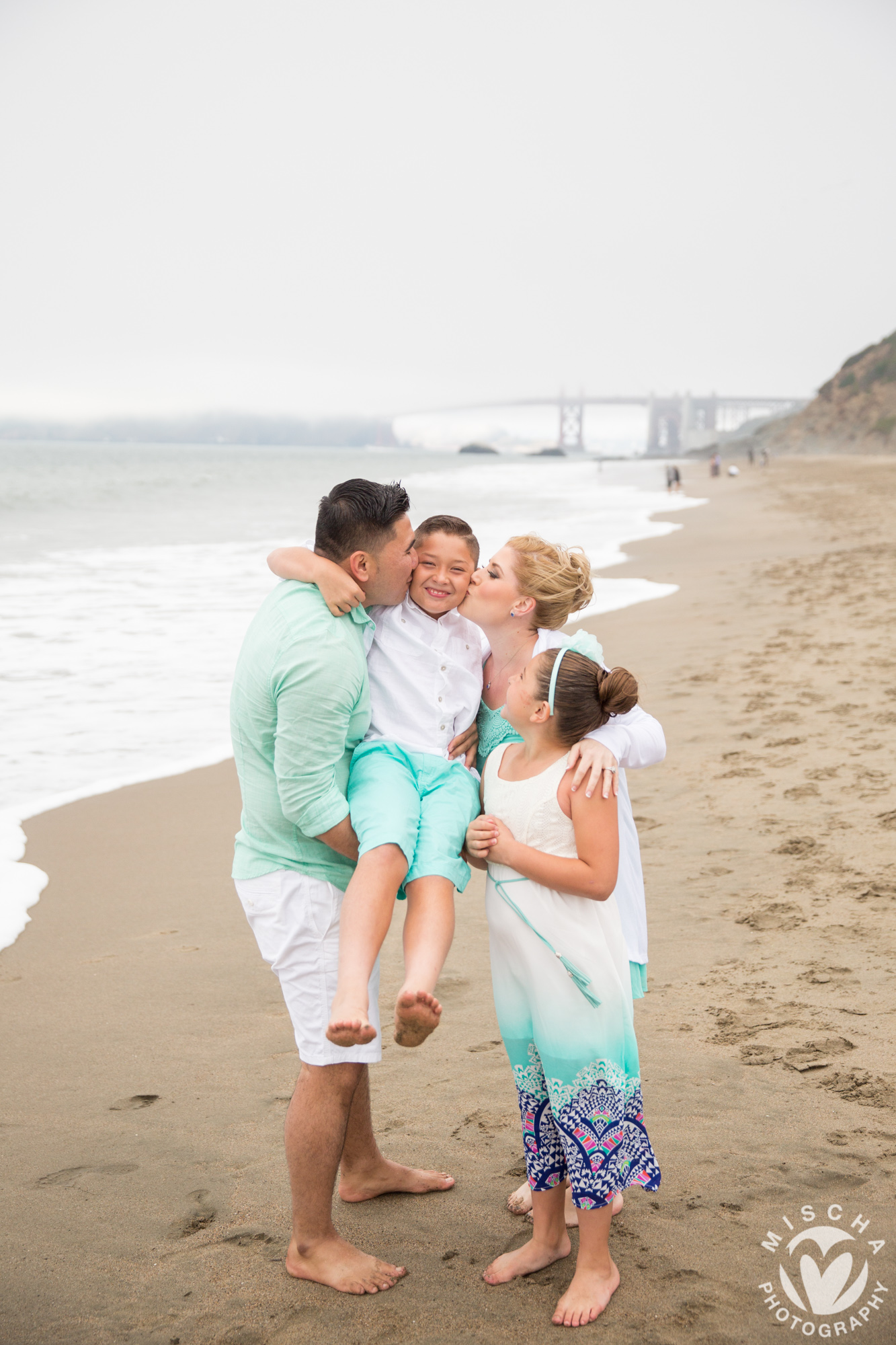 Family of four on beach with father holding boy, mother and daughter kissing boy, Golden Gate Bridge in background.