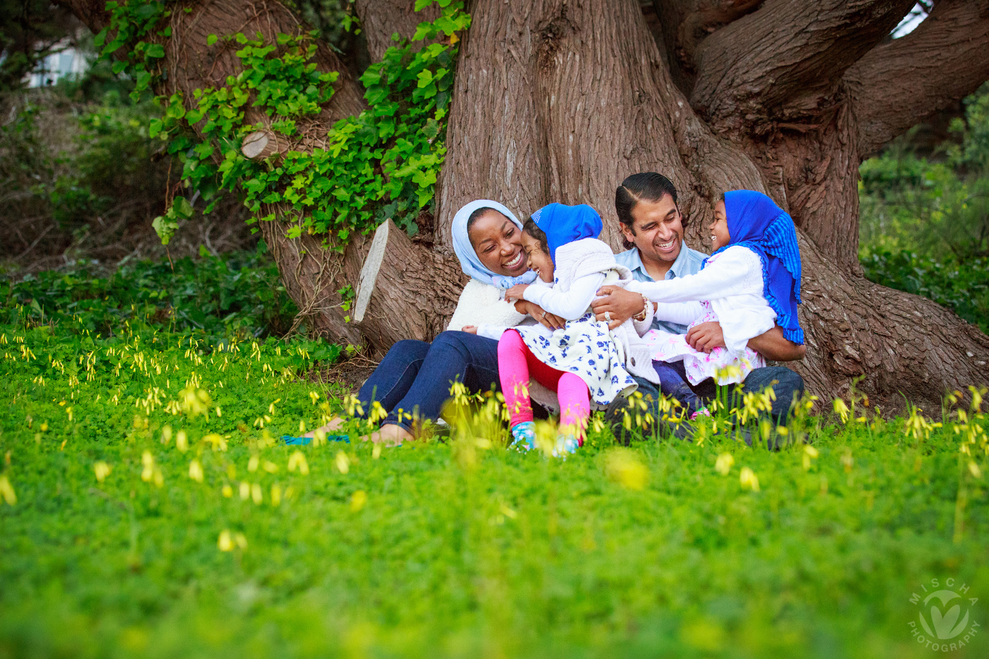 Family of four wearing hijabs sitting and smiling on grass near a large tree with yellow flowers.