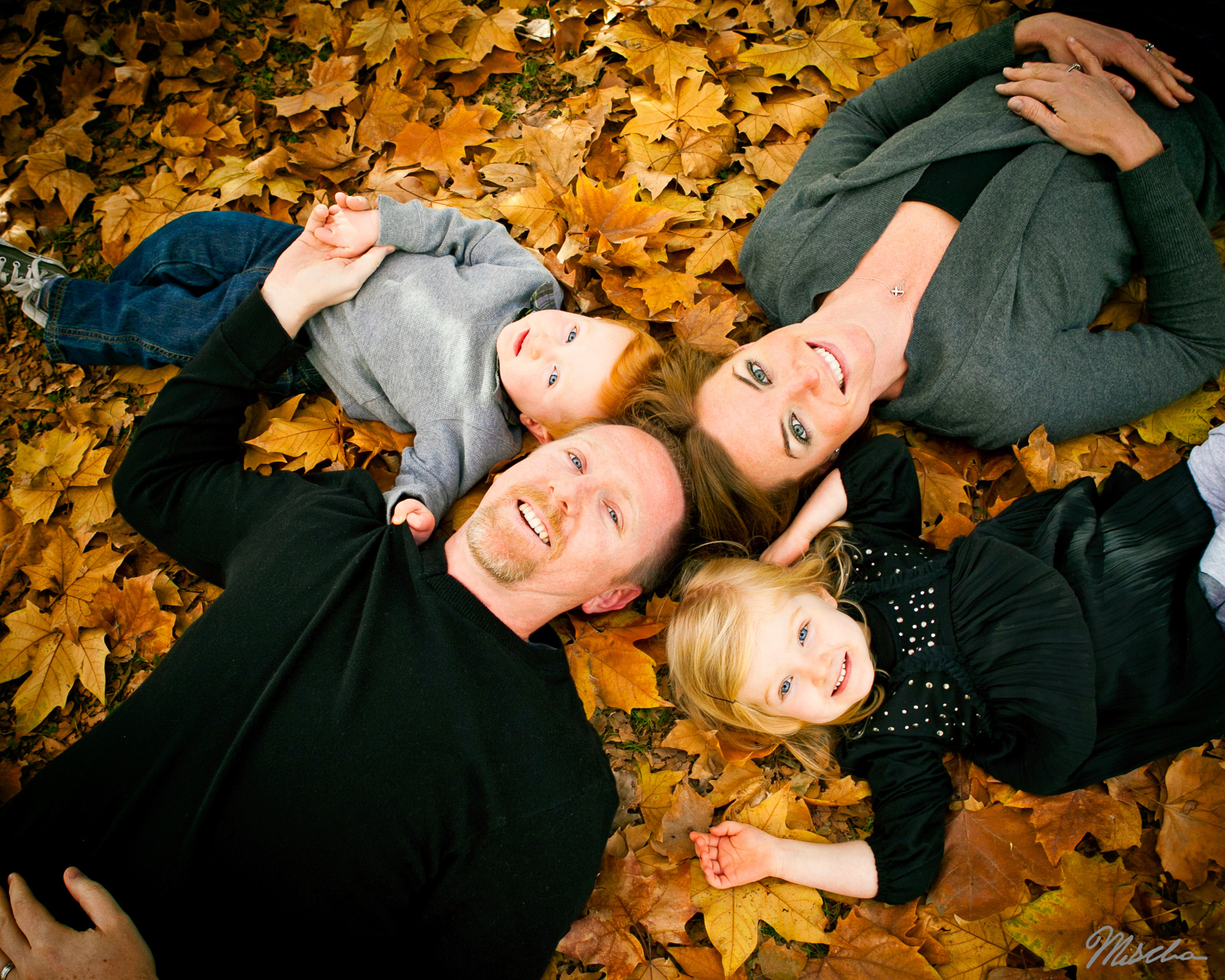 Family of two adults and two children lying on autumn leaves looking up at the camera.