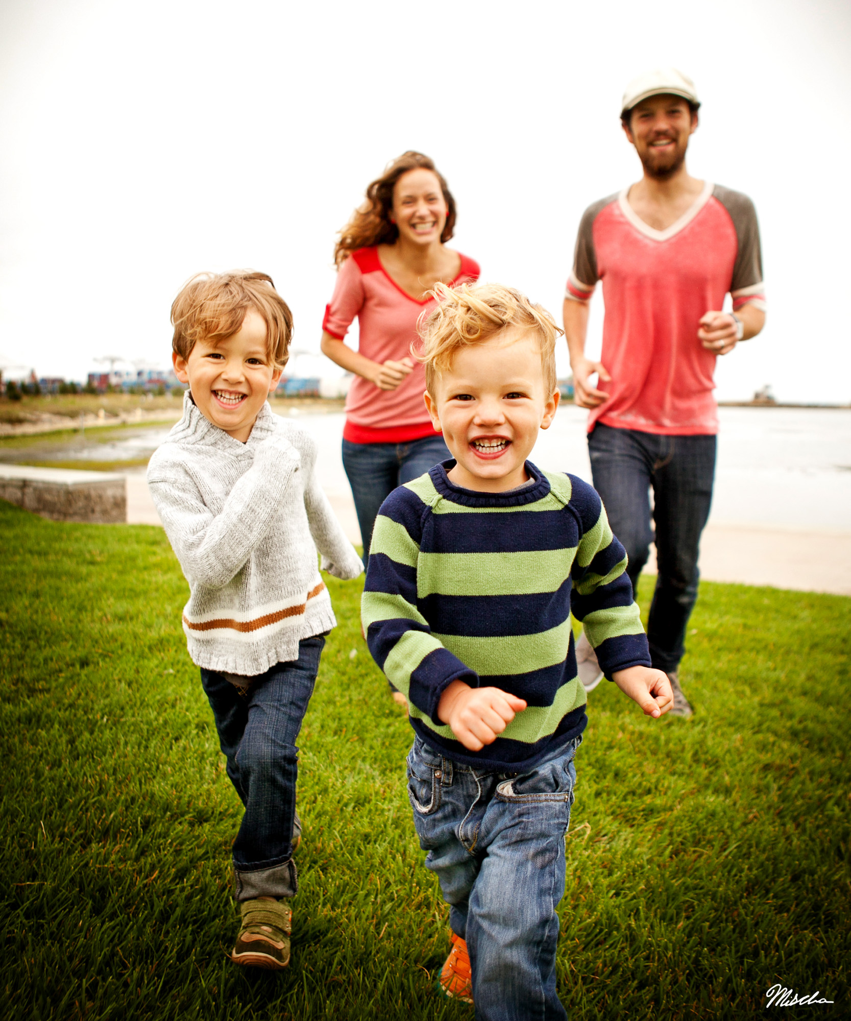 Two young boys running on grass with two adults jogging behind near water on a cloudy day