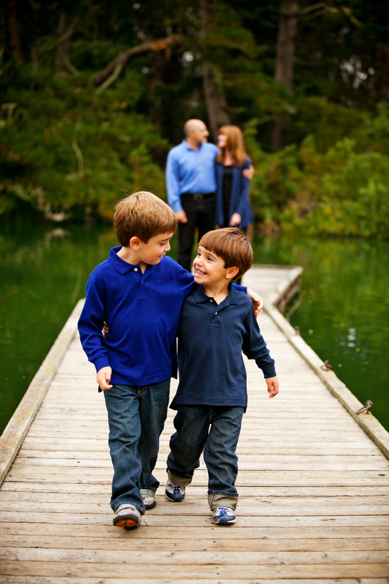 Two boys walk arm-in-arm on a wooden dock over water with trees and adults in background.