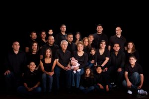 Multigenerational family portrait with 25 people wearing black tops and blue jeans on black background.