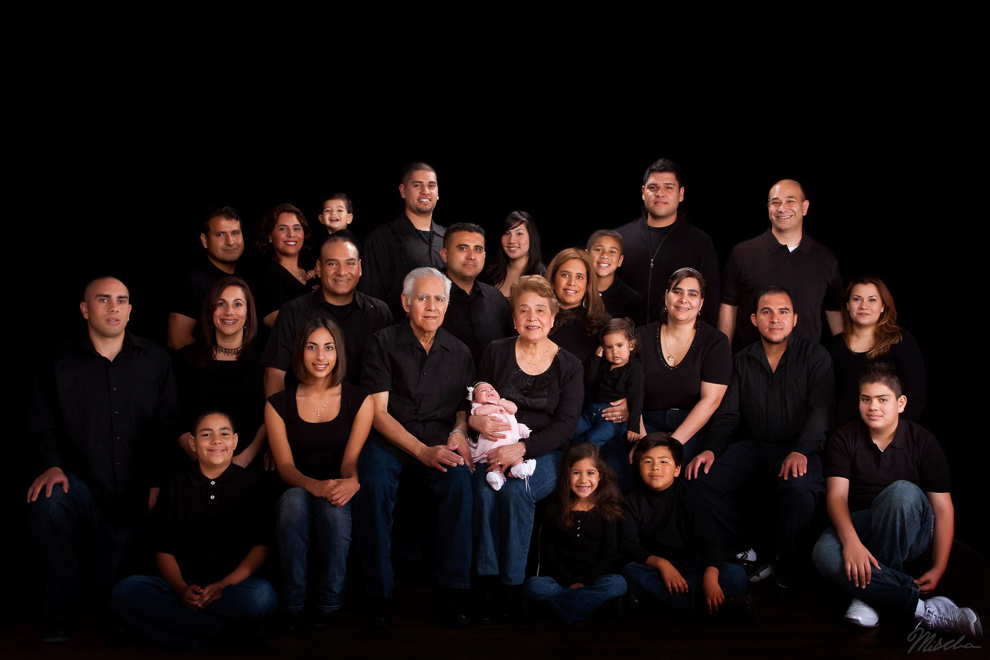 Multigenerational family portrait with 25 people wearing black tops and blue jeans on black background.