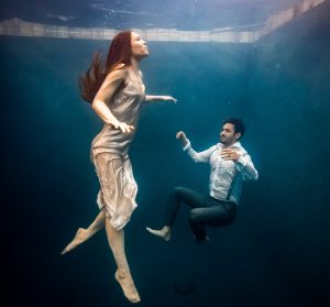 Underwater image of a woman in a dress ascending and a man in shirt and suspenders floating in a pool.