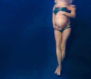 Pregnant woman underwater wearing dark bikini holding her belly against blue background.