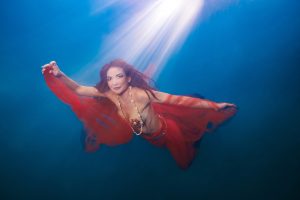 Underwater woman wearing red flowing skirt and shell necklace with light beam above.