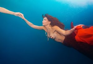 Underwater image of a topless woman with long hair wearing a shell necklace, holding a hand.