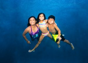 Three children in swimwear underwater holding each other against a blue water background