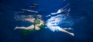 Underwater image of a pregnant woman in green and a man in white facing each other in blue water.