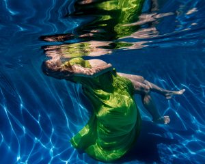 Pregnant woman in green dress floating underwater with hands on belly and eyes closed.