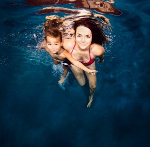 Woman in red bikini swimming underwater holding a young boy in a pool facing the camera