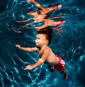 Young child swimming underwater in a pool wearing red swim trunks with light reflections.