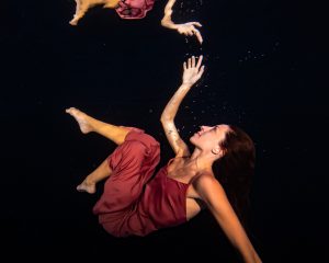 Woman in a red dress floating underwater with bubbles and her reflection above.