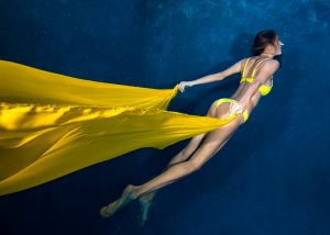 Woman in yellow bikini swimming underwater holding flowing yellow fabric trailing behind.