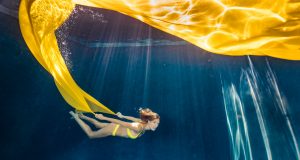 Woman in yellow bikini swimming underwater holding large flowing yellow fabric with light rays.
