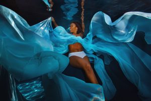 Person underwater wearing white bottoms with flowing blue fabric spreading around them.