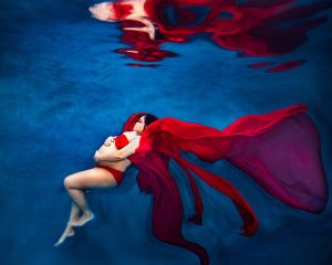 Pregnant woman in red bikini and flowing fabric floating underwater against blue background