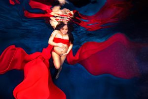 Pregnant woman underwater wearing flowing red fabric gently holding her belly.