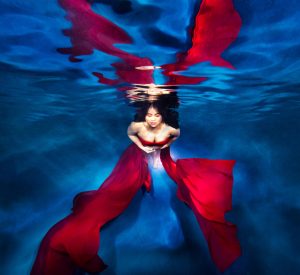 Woman underwater in a flowing red dress with light reflecting on the water surface above.